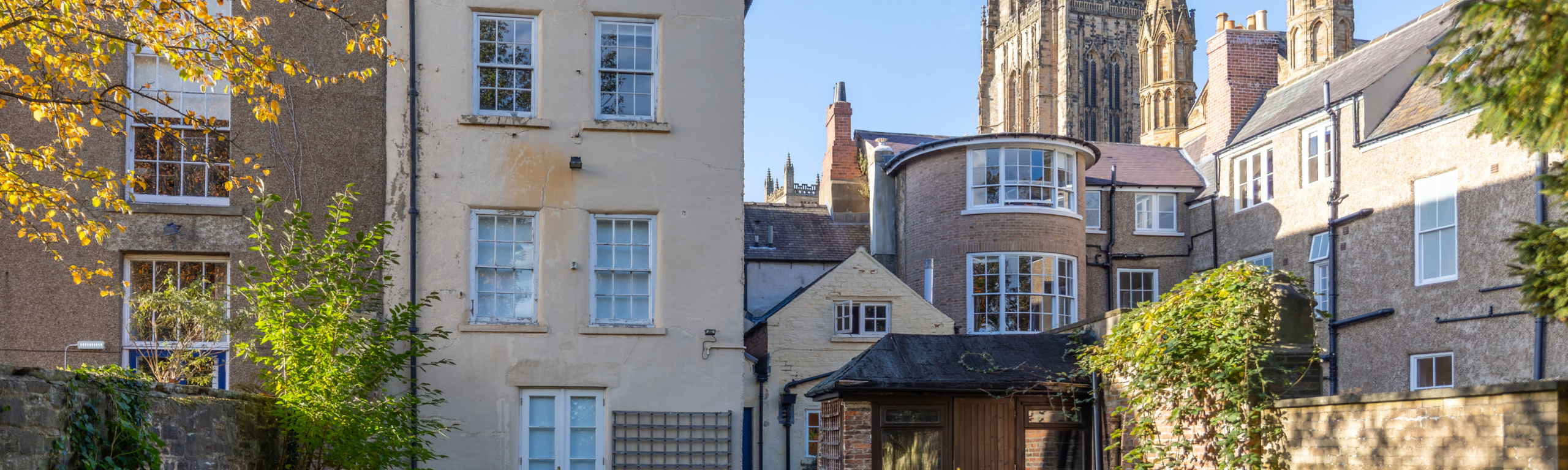 The rear courtyard of St John's College, featuring traditional multi-pane windows, climbing greenery, and a scenic view of Durham Cathedral towers in the distance