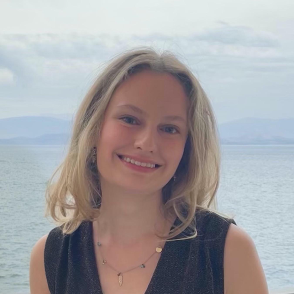 A young blonde woman smiles at the camera with the sea in the background